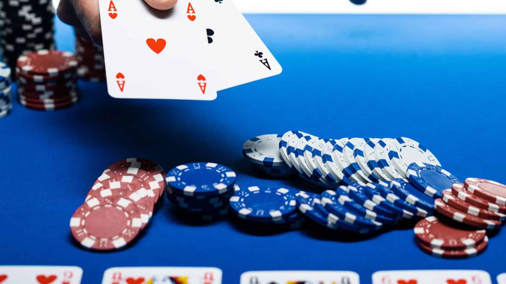 Igra? drži asa iznad stola prekrivenog plavim platnom, okružen gomilom poker ?ipova.