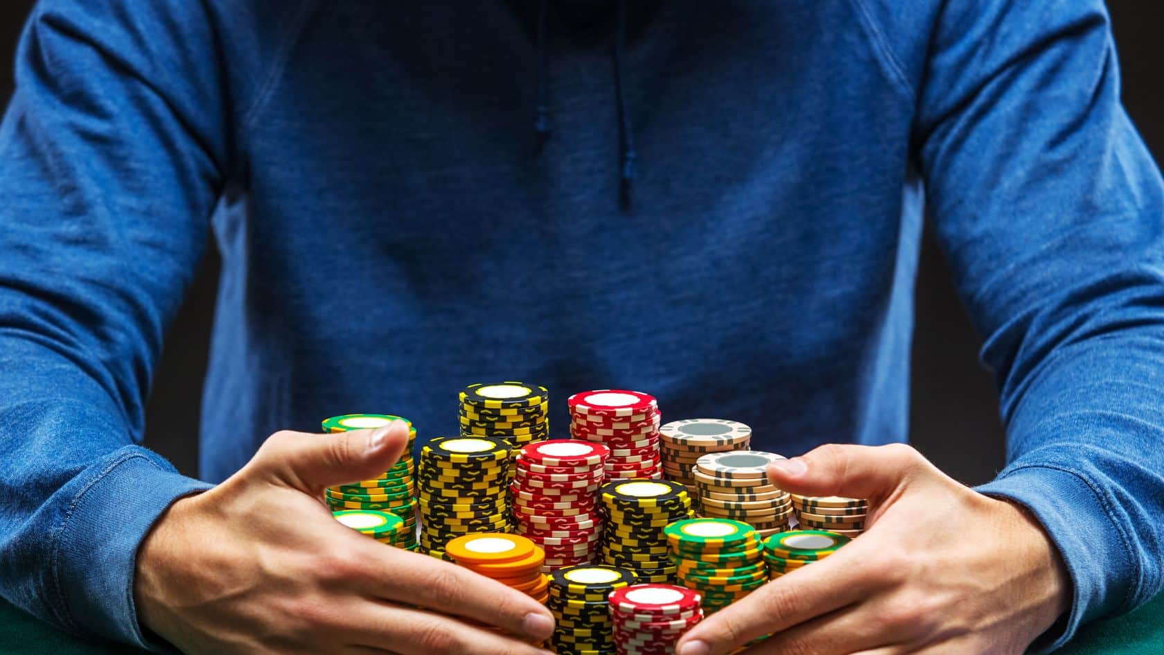 Muškarac u plavoj dukserici obuhvata veliku gomilu šarenih žetona na poker stolu, prikazuju?i visok ulog
