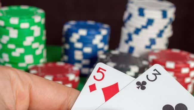  Igra? drži karte 5 karo i 2 tref, sa gomilom poker žetona u pozadini.