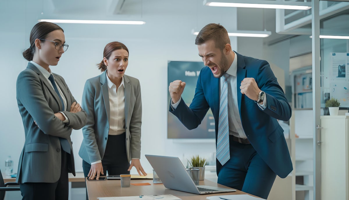 Muškarac u plavom odijelu slavi ispred laptopa u uredu, sa stisnuto pesnicama od uzbu?enja, dok dvije kolegice gledaju zbunjeno.