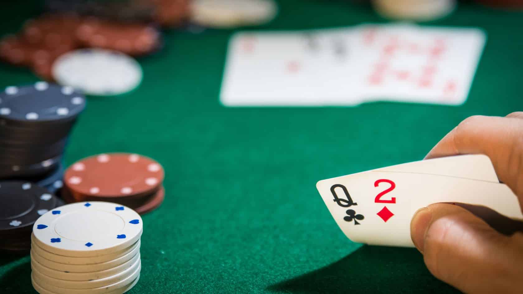 Ruka igrača otkriva kombinaciju dama tref i dvojka karo (Q♣ i 2♦) na zelenom poker stolu, uz gomile žetona u pozadini